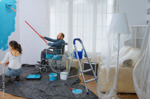 Obraz na płótnie Handicapped man in wheelchair helping wife to paint apartment wall, with handicapped, disabled ill and immobilizes man helping with apartment redecoration and home construction while renovating and