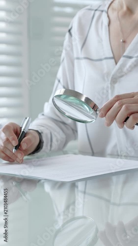 Woman's hands using a magnifying glass to scrutinize a document while holding a pen, researching important information for legal, financial, or academic analysis and validation