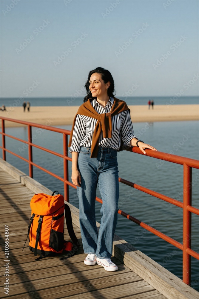 Fototapeta premium Woman with orange backpack stands on wooden pier. She smiles, enjoying sunny coastal weather. Casual outfit suits seaside travel and beach walks. Summer vacation vibes.