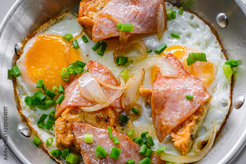 Close up of pan fried eggs topped with ham, sauteed onions, and chopped green onions served in a stainless steel skillet. Delicious Asian breakfast concept. Food close up.