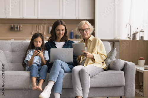 Tableau sur toile Absorbed senior 60s granny, her grown-up 30s daughter and pre-teen 10s granddaughter using diverse wireless devices seated on couch in living room