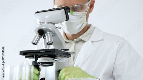 Scientist wearing a face mask, safety goggles, and protective gloves, precisely adjusting a microscope for microscopic examination and biological research in a lab