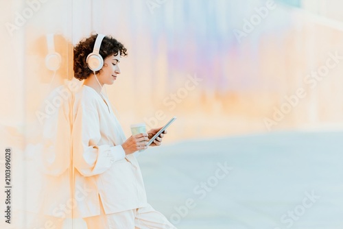 A young woman in cream scrubs relaxes against a city wall with a tablet and coffee, featuring a warm golden urban backdrop for healthcare break-time stock