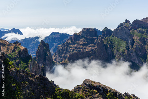 Dramatic mountain peaks rise above cloud sea. Pico do Areeiro landscape showcases rugged cliffs, lush greenery. Portugal's Madeira island offers breathtaking highland views.