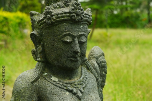 Ancient stone statue of a serene deity featuring intricate traditional carvings and mossy textures captured in a close up side profile shot set within a lush green tropical garden environment.
