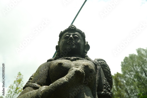 Antique stone statue of a serene goddess figure featuring intricate carvings and weathered textures captured from a low perspective against a bright sky in a lush tropical garden setting outdoors.