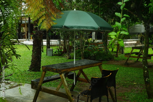 Wooden picnic table with a dark green umbrella sits in a lush garden setting during a rainy day featuring wet surfaces and natural foliage in a calm peaceful outdoor environment.