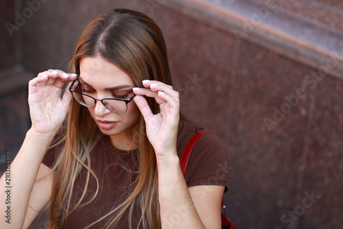 Close up portrait of a beautiful young woman with long light brown hair putting on eyeglasses while sitting near a granite wall in the city