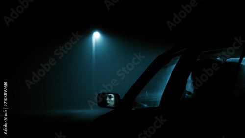 Woman looking in a rearview mirror of a car parked at night under a street light. Dark horror atmosphere with cinematic gloom and mystery. Concept of thriller scene for Halloween night.