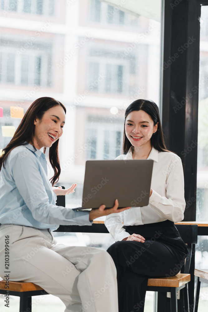 Obraz premium Two smiling Asian women collaborating using a laptop while sitting in a modern workspace