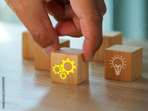 Wooden block with yellow gear icon held by hand among blocks including light bulb symbol, suggesting innovation and teamwork in business strategy and problem solving