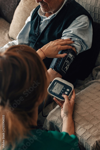 caregiver checks an elderly man's blood pressure at home. A monitor shows readings while both sit on a sofa in a warm, caring moment