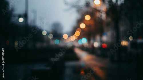 Softly defocused city street at twilight with glowing yellow and blue bokeh streetlights, blurred urban road and dark silhouettes of trees