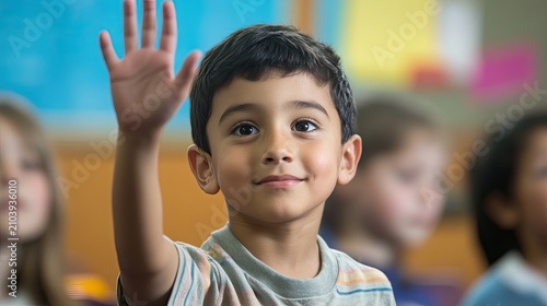 A young boy in a classroom raises his hand high, smiling at the camera. He is wearing a striped shirt and has dark hair.