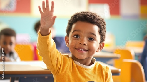 A young boy with curly black hair, wearing a yellow long-sleeve shirt, waves his hand in a classroom setting.
