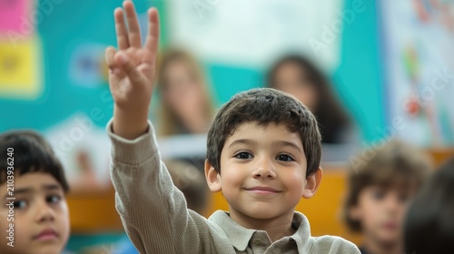 A young boy in a classroom raises his hand with three fingers, smiling at the camera.