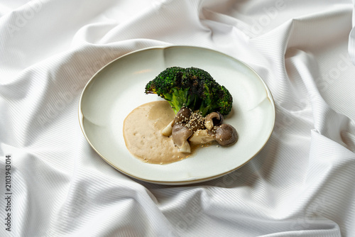 A plate features steamed broccoli and mushrooms served with a light sauce. The food is arranged neatly on a white tablecloth