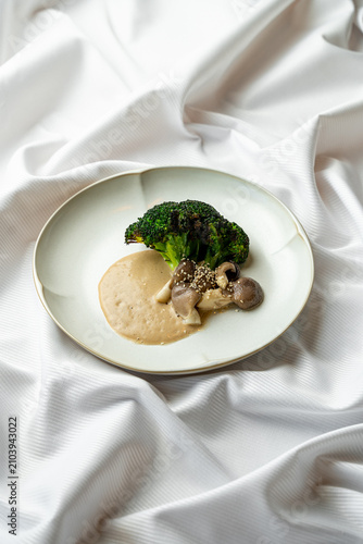 Broccoli sits on a white plate next to mushrooms and creamy sauce. The dish rests on a white tablecloth, showing a simple meal setup