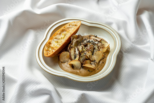 A creamy dish is served on a white plate alongside a slice of bread. The setting features a white cloth table covering. The time appears to be around lunchtime