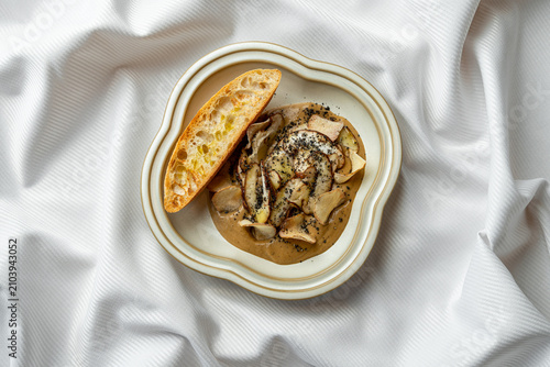 Pasta with mushrooms and a side of bread sits on a white plate. The dish is served on a plain white table, creating a simple meal scene
