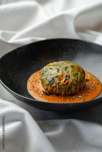 A round dish placed on a dark plate shows food surrounded by bright sauce. The setting has soft fabric in the background