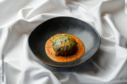 A dish is placed on a black plate. The food has a green topping and is surrounded by red sauce. The setting features a white cloth in the background