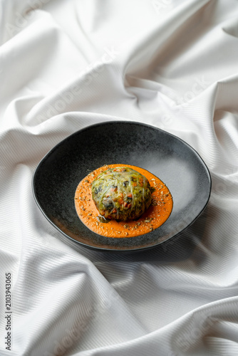 A round dish sits on a black plate. It has green filling wrapped in a thin layer. The plate is topped with orange sauce and some small garnishes. The background is white