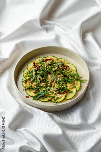 A plate holds a zucchini salad topped with nuts and herbs. The salad sits on a round dish against a smooth white background. It is arranged neatly and looks inviting