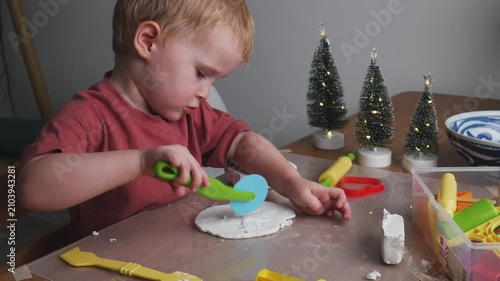 Toddler playing with air dry clay and making handmade holiday decorations at home. Three-year-old boy shaping air dry clay with toy tools at home. Cozy holiday atmosphere with miniature Christmas tree