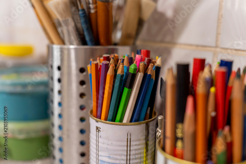 Colorful wooden pencils stored inside recycled tin cans on a workbench in a creative art studio or workshop workspace, closeup view with plastic paint containers nearby.