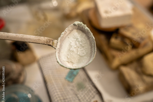 Konstfotografi Metal spoon filled with white enamel powder or flux for firing, held over a workshop table with refractory firebricks in the blurry background