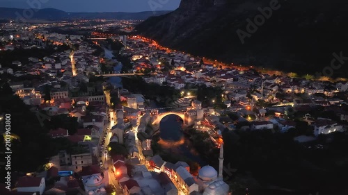 Mostar, Bosnia and Herzegovina - June 7, 2026: Drone flying left around the illuminated Old Bridge from the opposite riverbank