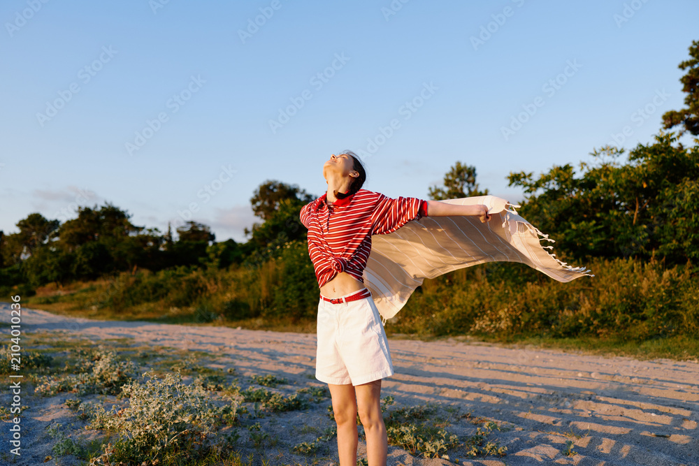 Fototapeta premium One woman wearing a striped red top and white shorts stands in a sunlit field, scarf flowing with the wind, expressing carefree summer fashion.