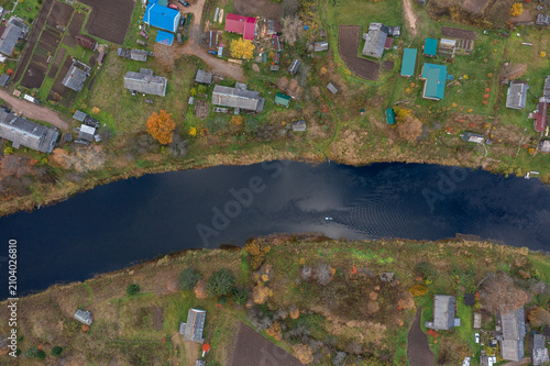 Fotografie Top-down aerial shot of a paddleboarder moving along a dark river dividing a rural village with neatly organized garden plots and colorful autumn trees