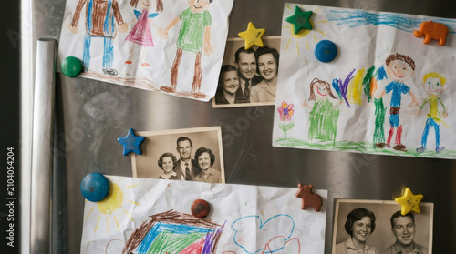 Children drawing and vintage family photo attached with magnet to metal refrigerator door. Home decor with nostalgic keepsakes representing childhood memories and intergenerational bond.