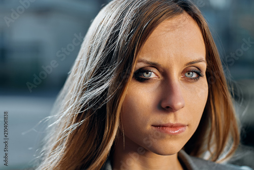 A close-up portrait of a serious young woman with striking blue eyes and long hair, photographed outdoors in natural light, conveying confidence, introspection and subtle professional elegance.