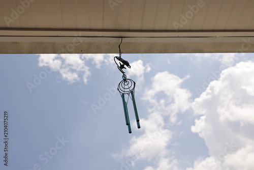 Decorative metallic wind chimes hang from a porch ceiling against a bright blue sky with clouds