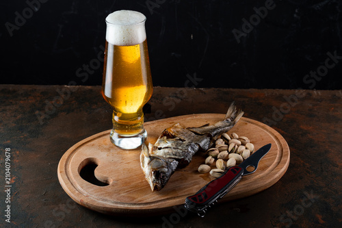 A glass of light lager beer with thick foam on a wooden cutting board with a folding pocket knife and sliced dried fish. Traditional beer snack still life concept on a rustic background.