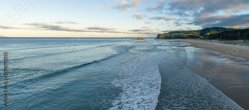 Sunrise over a serene beach in Opoutere, New Zealand. Gentle waves lap the shore as the sun begins to illuminate the sky, casting a warm glow on the landscape.
