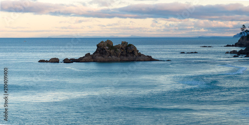 A small wave breaks near rocky outcrops in the ocean at sunrise. The sky is filled with soft clouds. Opoutere, New Zealand