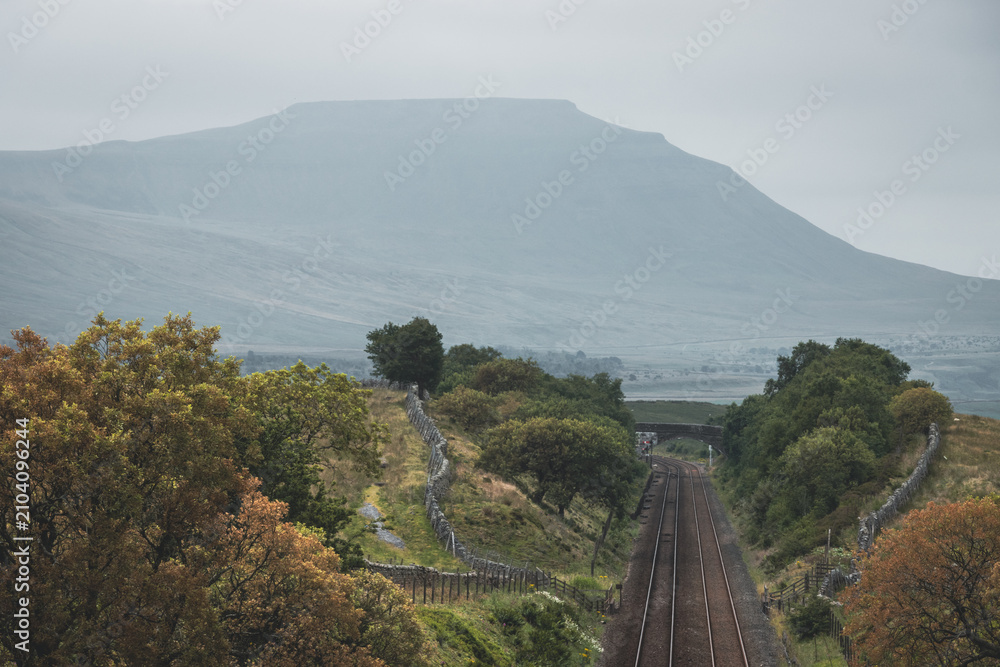 Obraz premium The historic Settle to Carlisle railway tracks pass under the stone Force Gill Aqueduct, with the distinct flat-topped plateau peak of Ingleborough rising into a misty, overcast Yorkshire sky.