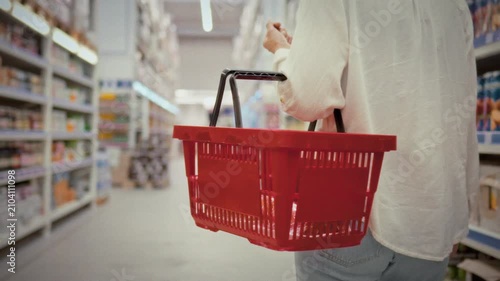 A girl strolls along the aisles of a modern supermarket with a red shopping basket in hand carefully looking at products on the shelves around her