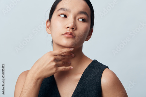 Foto Young woman with long black hair wearing dark sleeveless top posing and touching her neck with thoughtful expression on light gray background in studio portrait