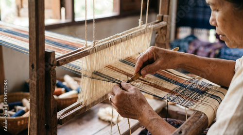 Close up of elderly artisan hands weaving traditional patterned fabric on wooden loom in workshop