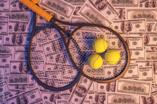 Fotografía Tennis rackets and balls lying on background of dollar banknotes in neon light showing sports betting and prize money in professional sport