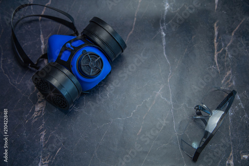 A blue respirator and goggles are spaced apart on a dark marble table. Concept, minimalism in labor protection, respiratory and visual protection, free space for text, safety. High quality photo