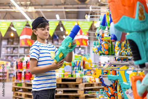 young boy holds a water gun while choosing toys in a modern retail store. Childhood shopping, summer play and family retail experience.