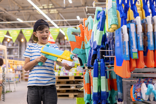 Happy little boy holding water blaster while choosing toy in modern toy store. Childhood shopping, summer play and family retail experience.