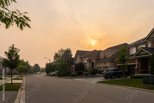 High-angle shot of a quiet suburban street with modern brick houses, set against an eerie orange and hazy sky caused by wildfire smoke obscuring the evening sun.