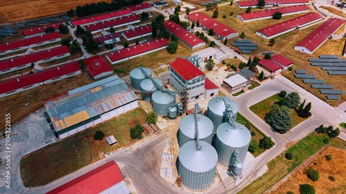 Grain storage facility within a farm complex. Oblique drone view of metal bins, conveyors, red roof barns, offices, paved roads and photovoltaic arrays.